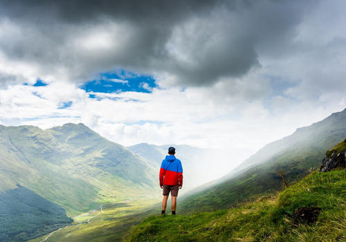 A male standing on a hill looking at mountains with the clouds coming around